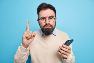 Strict serious bearded adult man raises index finger has excellent idea uses new mobile application holds smartphone wears spectacles and sweater isolated over blue backgound. Technology user