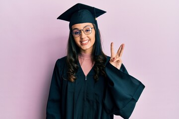 Young hispanic woman wearing graduation cap and ceremony robe showing and pointing up with fingers number three while smiling confident and happy.