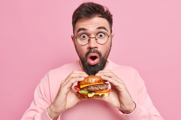 Stupefied handsome man poses with delicious appetizing fast food holds tasty hamburger wears round spectacles casual jumper doesnt keep to diet likes cheat meal dressed casually isolated on pink wall