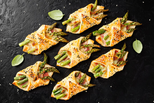 Baked Green Asparagus With Ham And Cheese In Puff Pastry Sprinkled With Sesame Seeds And Green Basil Leaves. Black Stone Background, Top View