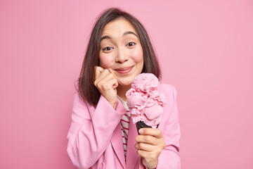 Attractive brunette young Asian woman with delighted expression holds tasty cone ice cream in waffle keeps hand on face has sweet tooth isolated over pink studio background. Yummy summer dessert