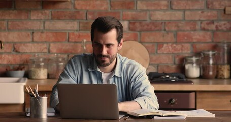 Serious millennial man sitting at table in kitchen check written info at personal organizer with recording information, working at home, texting on laptop device. Telecommute, e-learn activity concept