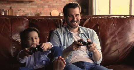 Young father and little 7s son sit on couch enjoy videogames, lively cheerful dad and small child boy holding game pads having fun on weekend at home. Hobby, playtime and leisure with kids concept