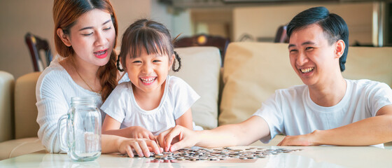 Banner photo of Asian family teach their daughter to saving money and putting coins into glass bank spend a quality time together. Asian family, relationship, happiness and investment concept