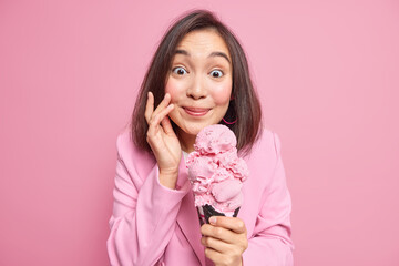 Summer is here. Pretty lovely brunette young eastern woman holds big strawberry ice cream in waffle enjoys eating delicious frozen dessert wears fashionable jacket isolated over pink background