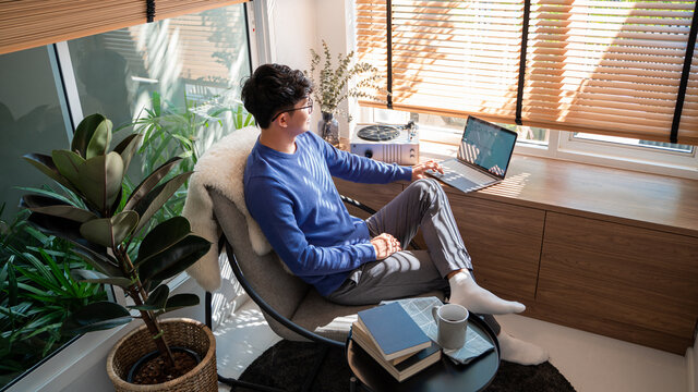 Young asian male tech user relaxing  holding laptop computer and looking at the screen in living room, Remote Job or work from home concept.