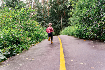 little girl in yellow rubber boots walks along the road in the park on a summer day