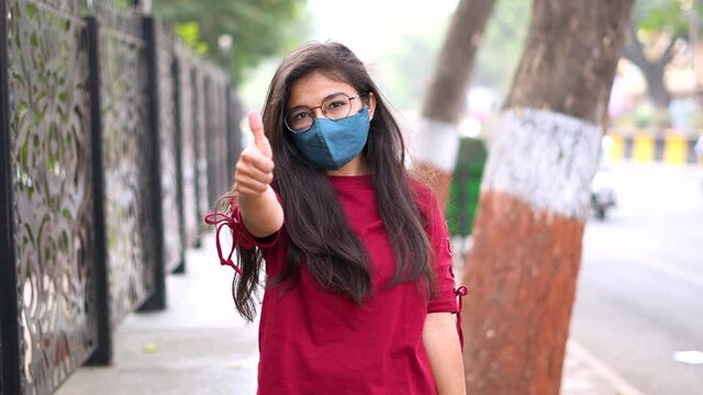 Portrait of young Indian girl in stylish mask and giving thumbs up while standing on street of city during corona virus pandemic