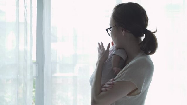 portrait of happy asian woman mother holding sleepy baby infant near window at home daytime. 4 months baby embracing in parent arm. relationship mother and daughter. mom touching baby lull to sleep.