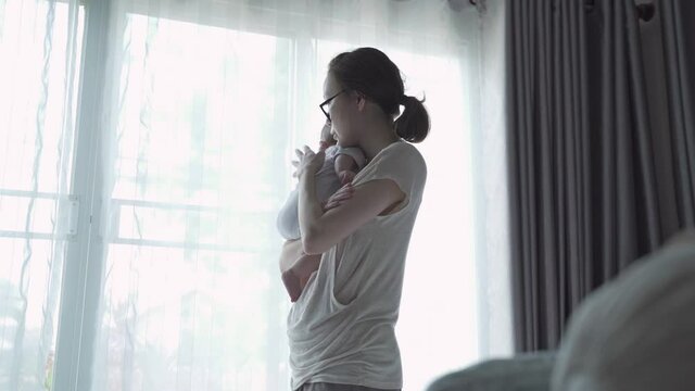 portrait of happy asian woman mother holding sleepy baby infant near window at home daytime. 4 months baby embracing in parent arm. relationship mother and daughter. mom touching baby lull to sleep.