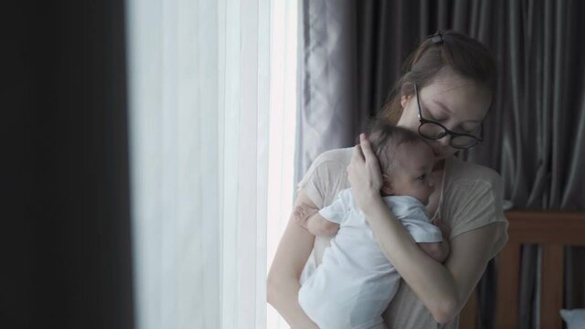portrait of happy asian woman mother holding sleepy baby infant near window at home daytime. 4 months baby embracing in parent arm. relationship mother and daughter. mom touching baby lull to sleep.