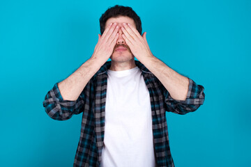 young Caucasian man with moustache wearing plaid shirt against blue wall covering eyes with both hands, doesn't want to see anything or feeling ashamed. Human feelings reactions.
