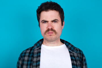 Fototapeta premium young Caucasian man with moustache wearing plaid shirt against blue wall frowning his eyebrows being displeased with something.