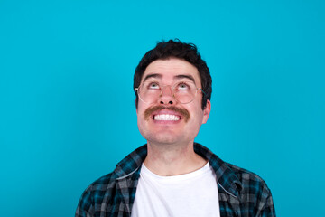 Portrait of mysterious young Caucasian man with moustache wearing plaid shirt against blue wall looking up with enigmatic smile. Advertisement concept.