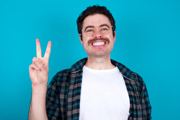 young Caucasian man with moustache wearing plaid shirt against blue wall showing and pointing up with fingers number two while smiling confident and happy.