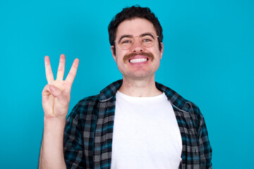 Naklejka premium young Caucasian man with moustache wearing plaid shirt against blue wall showing and pointing up with fingers number three while smiling confident and happy.