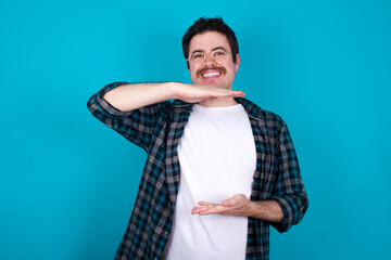 young Caucasian man with moustache wearing plaid shirt against blue wall gesturing with hands showing big and large size sign, measure symbol.