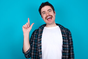 Pleasant looking young Caucasian man with moustache wearing plaid shirt against blue wall has...