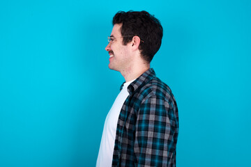 Profile of smiling young Caucasian man with moustache wearing plaid shirt against blue wall with healthy skin, has contemplative expression, ready to have outdoor walk.