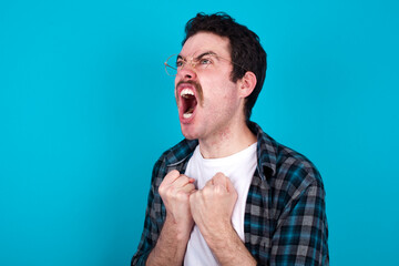 young Caucasian man with moustache wearing plaid shirt against blue wall excited and glad to achieve victory, clenches fists, screams in excitement with closed eyes,successful person.