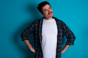 Funny frustrated young Caucasian man with moustache wearing plaid shirt against blue wall holding hands on waist and silly looking at awkward situation.