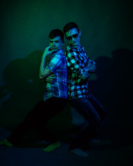 Fototapeta na wymiar Two young men in plaid shirts dance in the studio with multicolored light