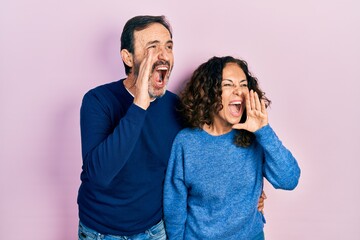 Middle age couple of hispanic woman and man hugging and standing together shouting and screaming loud to side with hand on mouth. communication concept.