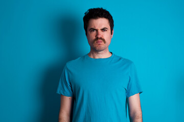Offended dissatisfied young handsome Caucasian man with moustache wearing blue t-shirt against blue background with moody displeased expression at camera being disappointed by something