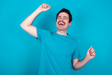 Photo of upbeat young handsome Caucasian man with moustache wearing blue t-shirt against blue has fun and dances carefree wear being in perfect mood makes movements. Spends free time on disco party