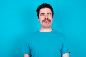 Funny young handsome Caucasian man with moustache wearing blue t-shirt against blue background makes grimace and crosses eyes plays fool has fun alone sticks out tongue.