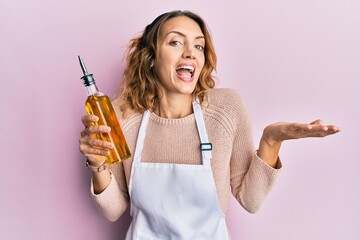 Young caucasian woman wearing apron holding olive oil can celebrating achievement with happy smile and winner expression with raised hand
