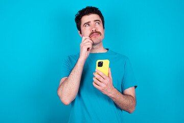 young handsome Caucasian man with moustache wearing blue t-shirt against blue wall thinks deeply...
