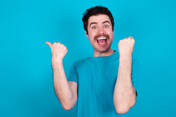 Hooray cool young handsome Caucasian man with moustache wearing blue t-shirt against blue background point back empty space hand fist