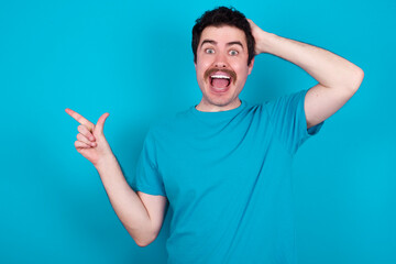 Fototapeta premium Surprised young handsome Caucasian man with moustache wearing blue t-shirt against blue background pointing at empty space holding hand on head