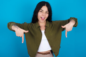 young beautiful tattooed girl standing against blue wall with positive expression, points down with both index fingers, has broad interested smile. Look there, please.