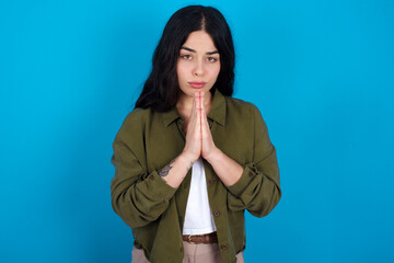 young beautiful tattooed girl standing against blue wall keeps palms pressed together in front of...