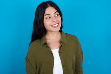 Oops! Portrait of young beautiful tattooed girl standing against blue wall clenches teeth and looks confusedly aside, realizes her bad mistake,