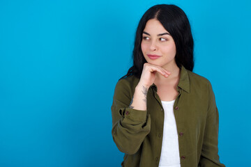 Dreamy young beautiful tattooed girl standing against blue wall with pleasant expression, looks sideways, keeps hand under chin, thinks about something pleasant.