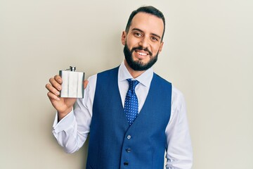 Young man with beard drinking whiskey from flask looking positive and happy standing and smiling with a confident smile showing teeth