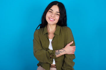 People, lifestyle, youth and happiness concept. Shy pretty young beautiful tattooed girl standing against blue wall, feeling happy hugging herself.