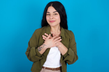 Honest young beautiful tattooed girl standing against blue background keeps hands on chest, touched by compliment or makes promise, looks at camera with great pleasure.