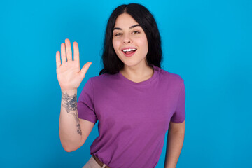 young beautiful tattooed girl wearing purple t-shirt standing against blue background waiving...