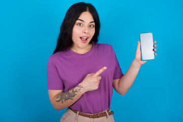 Excited young beautiful tattooed girl wearing purple t-shirt standing against blue background holding and pointing with finger at smartphone with blank screen. Advertisement concept.