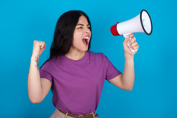 young beautiful tattooed girl wearing purple t-shirt standing against blue wall communicates shouting loud holding a megaphone, expressing success and positive concept, idea for marketing or sales.