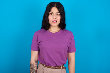 Portrait of dissatisfied young beautiful tattooed girl wearing purple t-shirt standing against blue smirks face, purses lips and looks with annoyance at camera, discontent hearing something unpleasant
