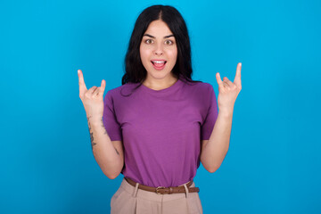 young beautiful tattooed girl wearing purple t-shirt standing against blue background makes rock n roll sign looks self confident and cheerful enjoys cool music at party. Body language concept.