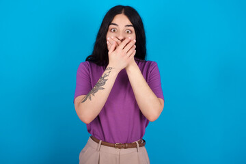 Stunned young beautiful tattooed girl wearing blue t-shirt standing against blue background covers mouth with both hands being afraid from something or after hearing stunning gossips.