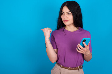 young beautiful tattooed girl wearing blue t-shirt standing against blue background points thumb away and shows blank space aside, holds mobile phone for sending text messages.