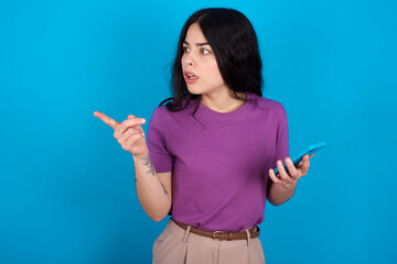 Stunned young beautiful tattooed girl wearing blue t-shirt standing against blue background points sideways right copy space, recommends product, sees astonishing thing
