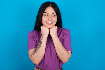 young beautiful tattooed girl wearing blue t-shirt standing against blue background holds hands under chin, glad to hear heartwarming words from stranger